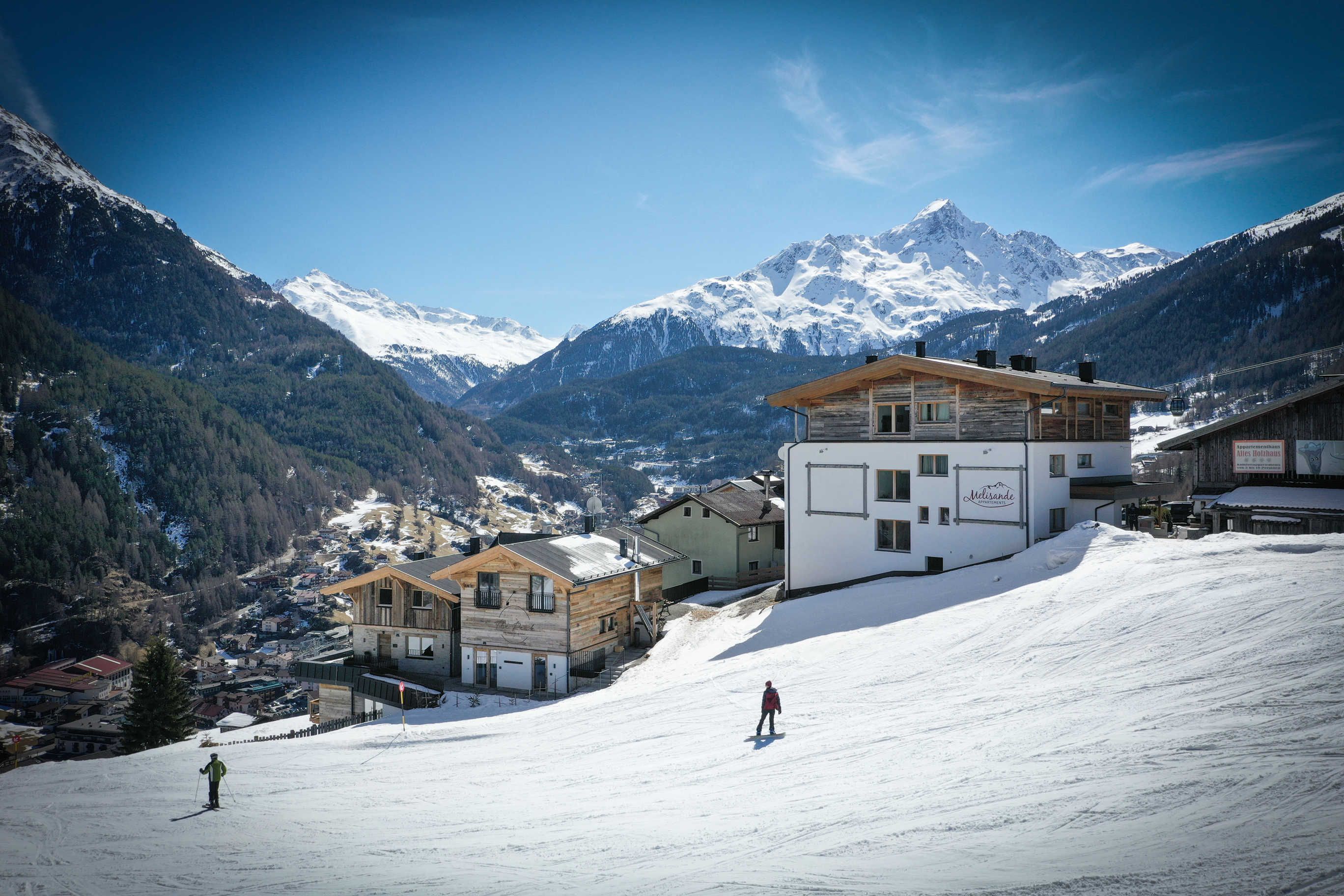 Een skigebied met houten gebouwen, een skiënd persoon en besneeuwde bergen op de achtergrond.