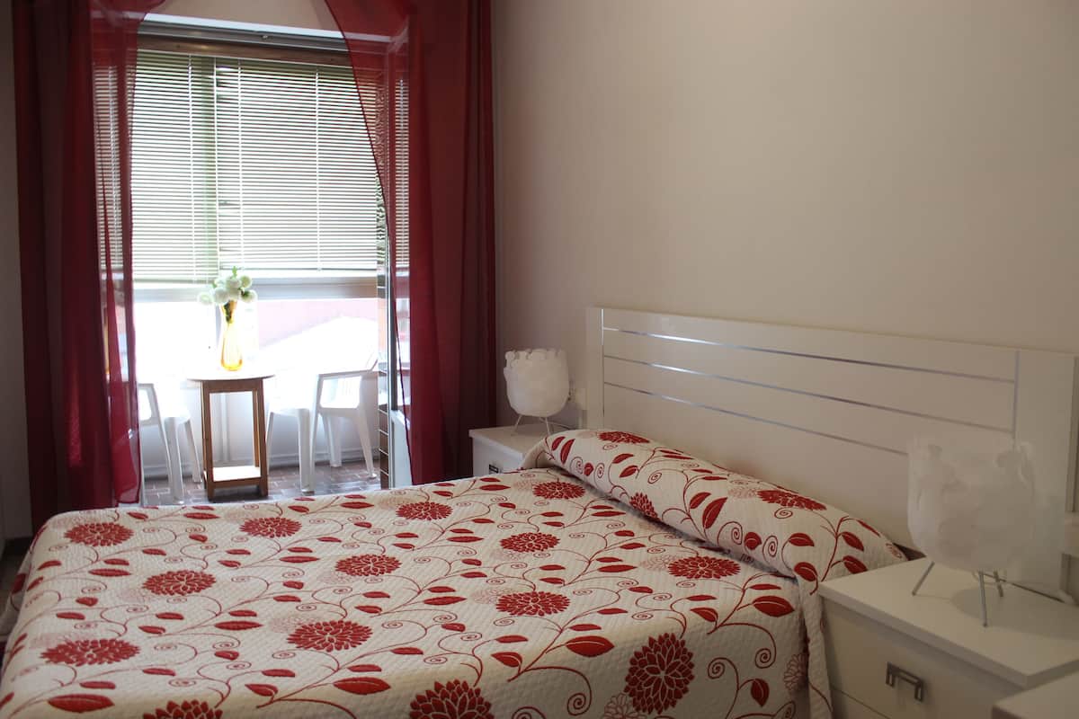A bedroom with a bed, a bedside table, a nightstand, a window with curtains, and a view of an outdoor area with a table and chairs.