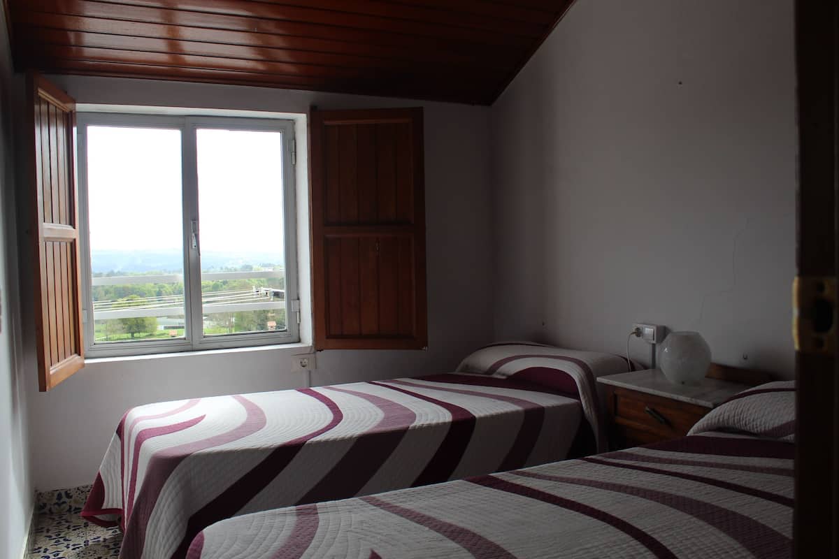 A bedroom with two beds, a window with a view, wooden ceiling, and a nightstand with a lamp.