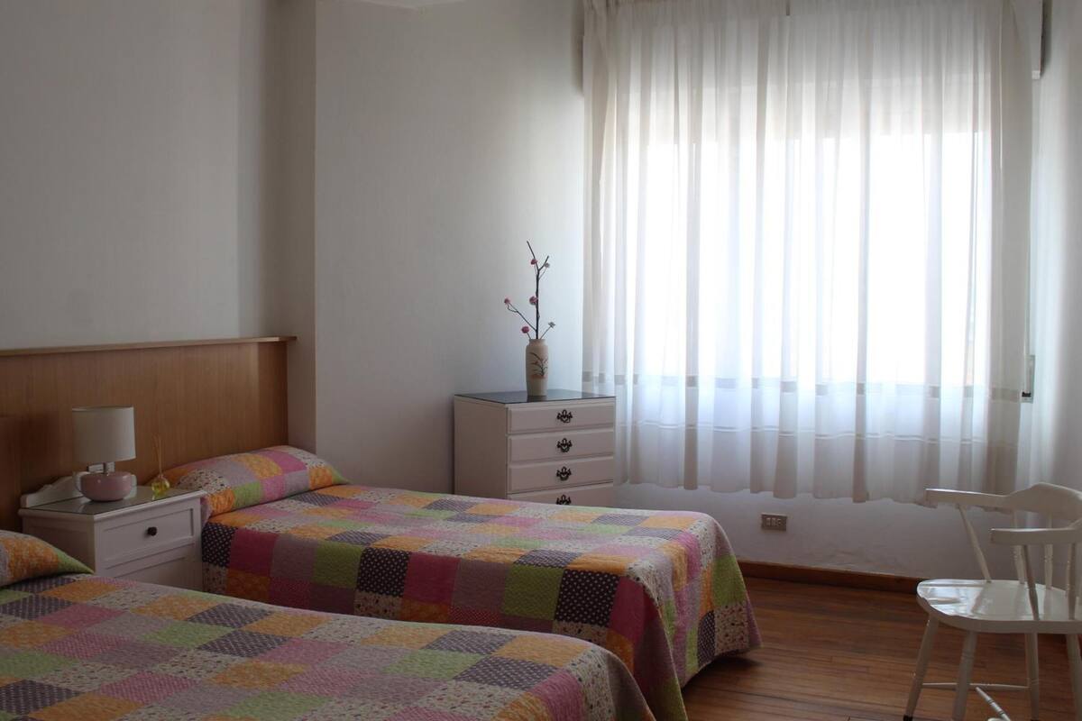 A bedroom with two beds, a nightstand, a dresser, a chair, and a window with sheer curtains.