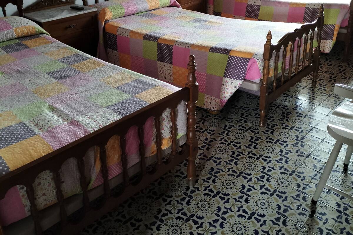 A room with three wooden beds, each with a colorful quilt. The room has patterned floor tiles and a window with natural light.