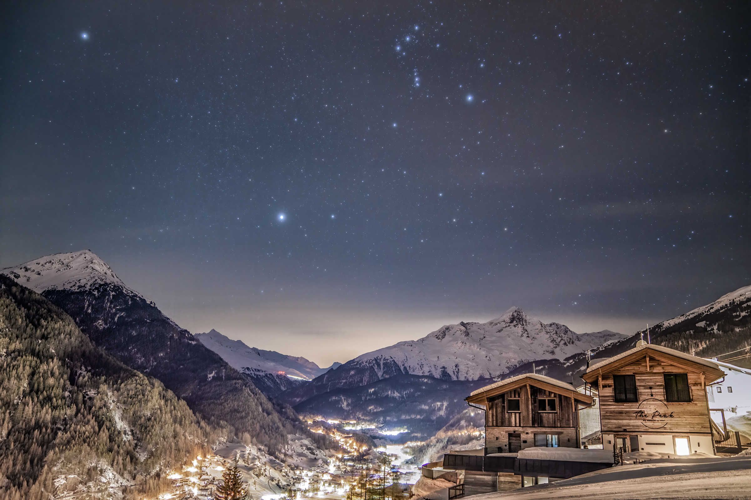 A mountain lodge with a starry night sky, snow-covered peaks, and a valley with lights.