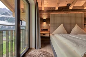 A bedroom with a bed, a nightstand, a wall-mounted light, a window with a view of mountains, and a wooden ceiling. - Bergsteiger Chalet - The Peak Sölden (Sölden)