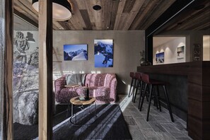 A modern interior with a wooden ceiling, a pink patterned sofa, a small round table, and a bar area with stools. - Bergsteiger Chalet - The Peak Sölden (Sölden)