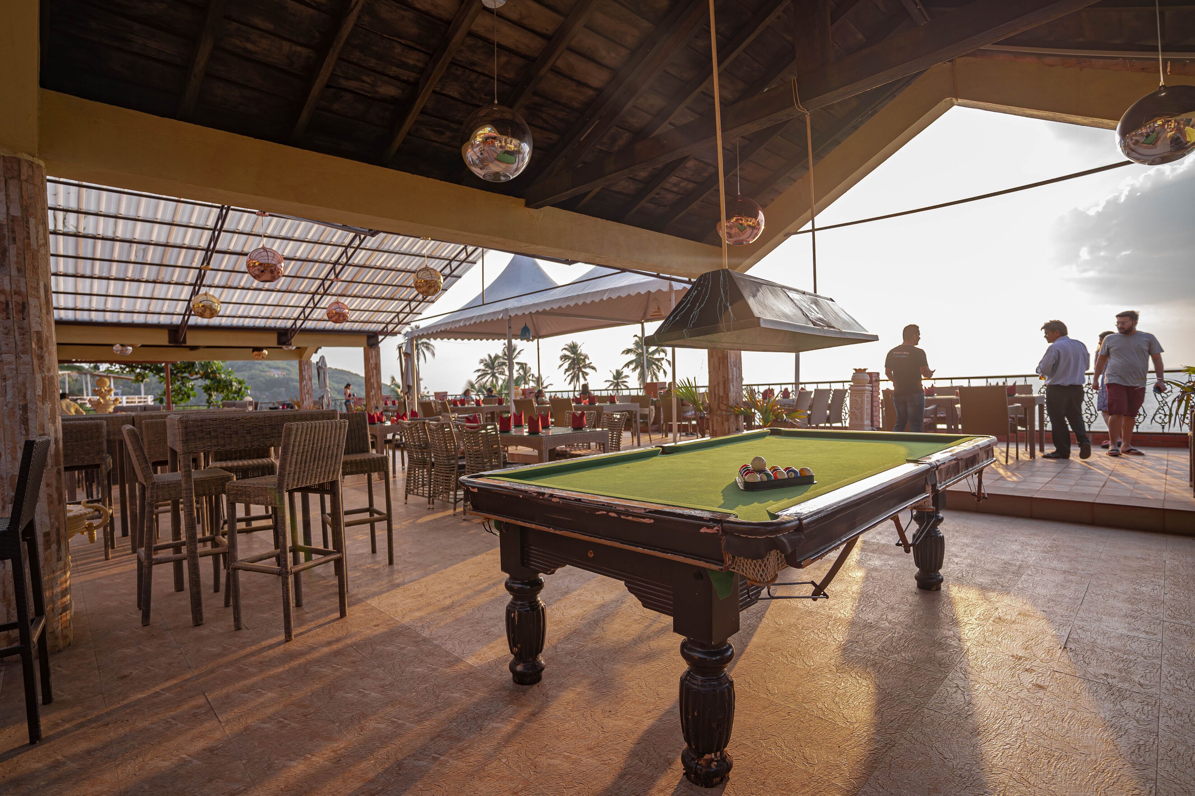 A pool table in a covered outdoor area with chairs and tables, and a view of palm trees and mountains in the background.