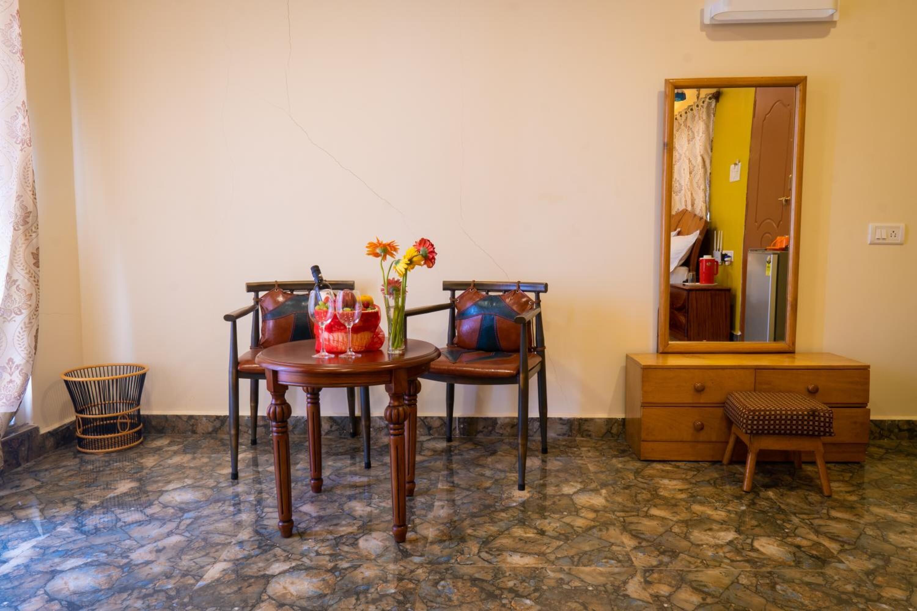 Ein Zimmer mit Steinboden, einem Holztisch mit Stühlen, einem kleinen Hocker, einer Holzkommode und einem Spiegel an der Wand.