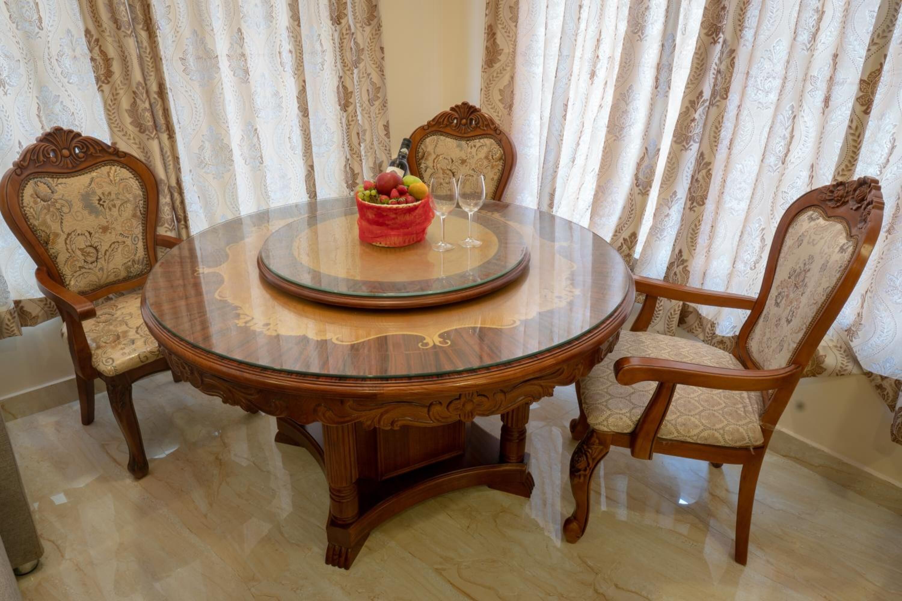 A round wooden dining table with a glass top, surrounded by two upholstered chairs with intricate designs.