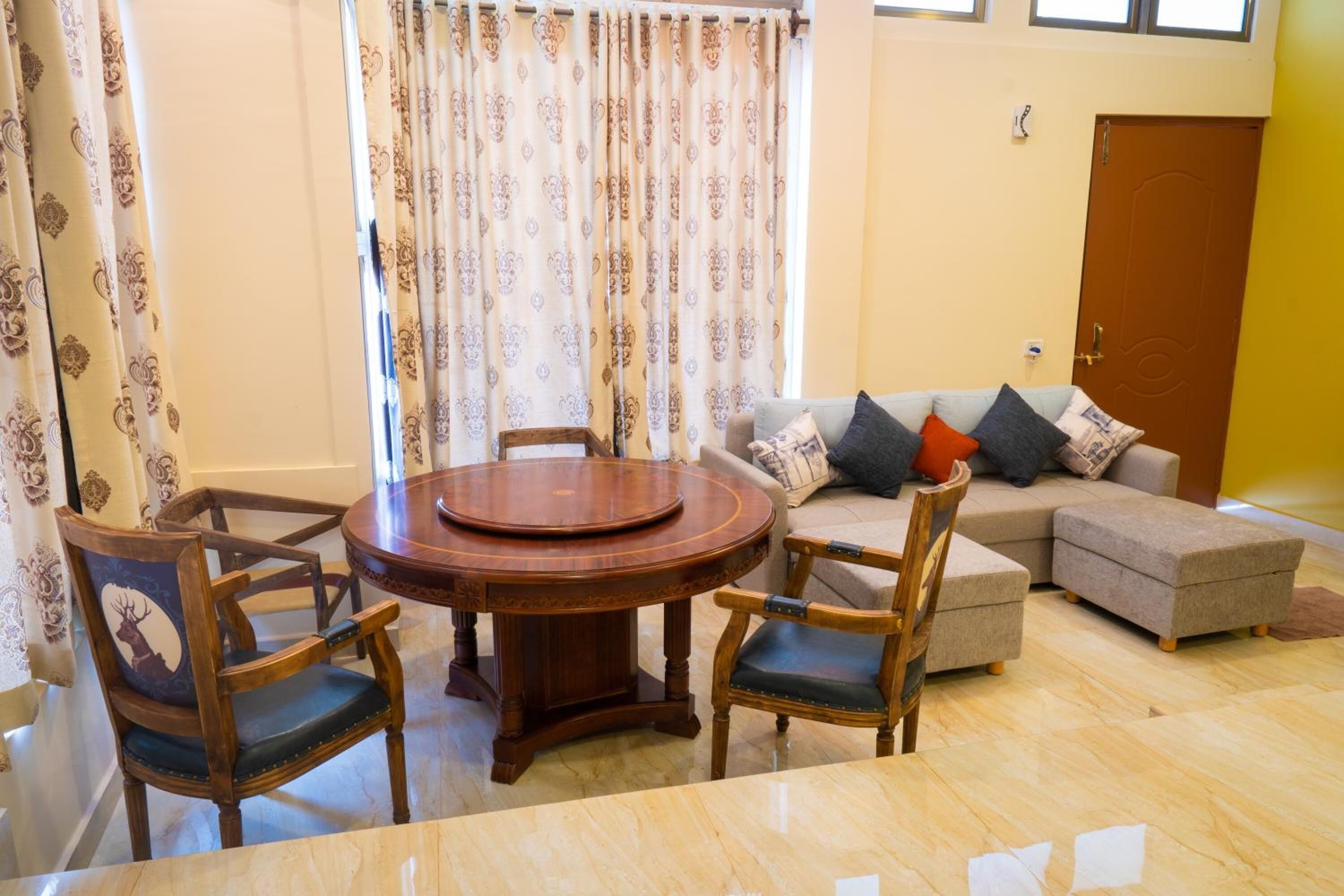 Een woonkamer met een ronde houten tafel, twee houten stoelen, een bank met kussens en een kamerkleed met patroon.