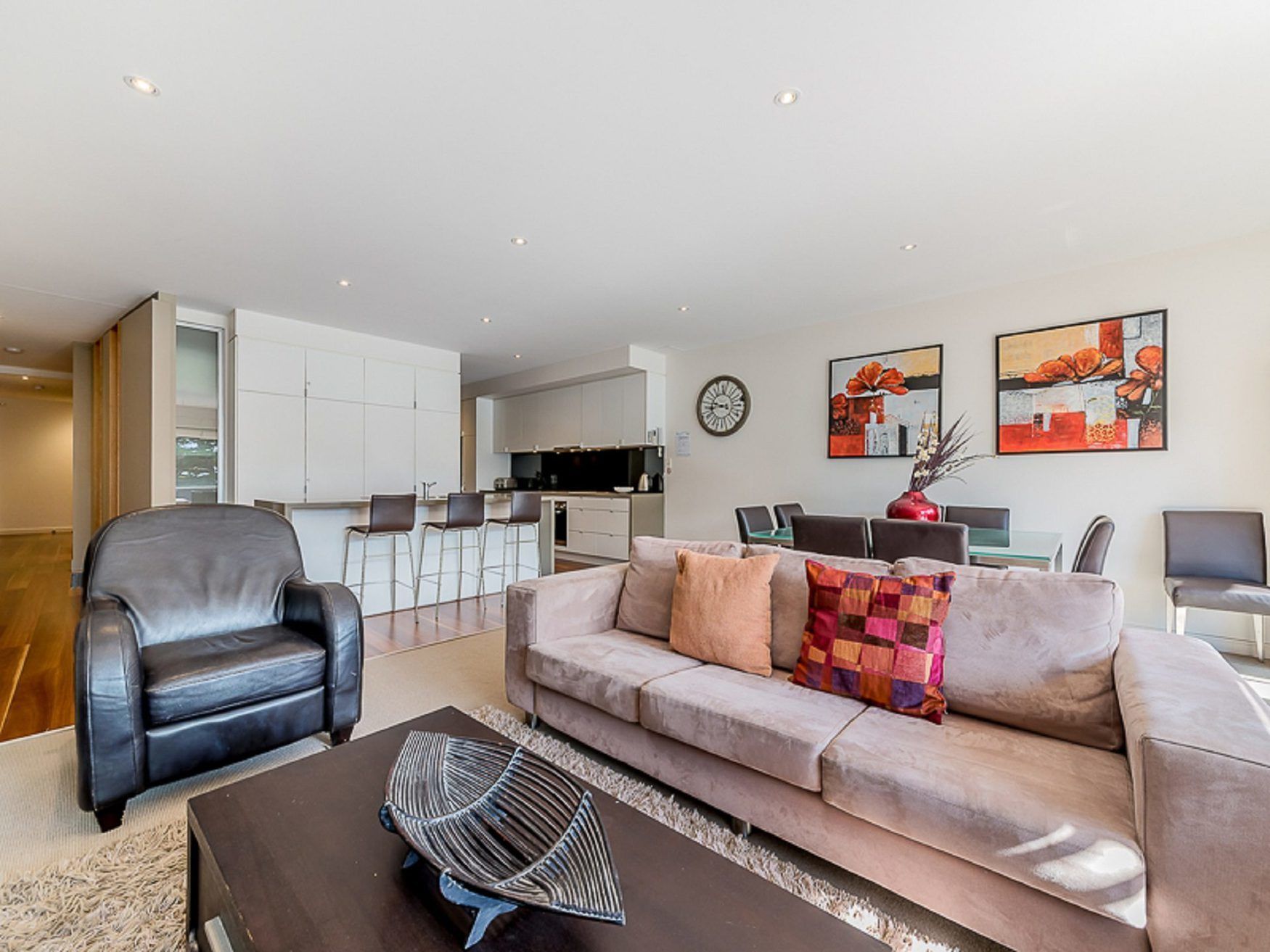 Un salon moderne avec un fauteuil en cuir, un canapé beige et une table basse. Au fond, on aperçoit une cuisine avec des meubles blancs et un coin repas comprenant une table et des chaises.