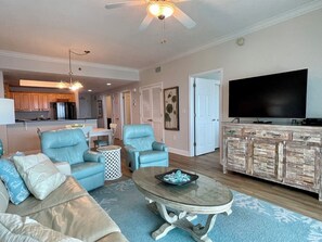 A living room with a sofa, chairs, a coffee table, a TV, and a kitchen area in the background.