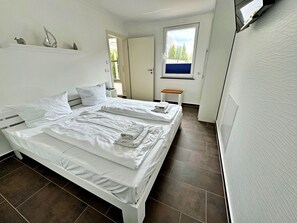 A clean, modern bedroom with a double bed, a small bedside table, and a window with a view of greenery. - Apartment Altenberg 02 - Apartment 02 (Heringsdorf)