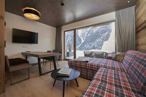 A modern living room with a flat-screen TV, a sofa, a dining table, and a view of mountains through large windows.