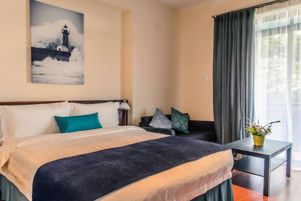 A hotel room with a bed, a nightstand, a lamp, a chair, a table with a plant, and a painting of a lighthouse on the wall.