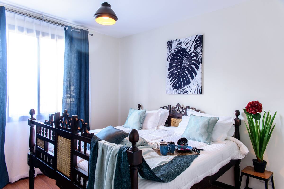 A neatly made bed with a wooden headboard, a potted plant, and a wall-mounted artwork.