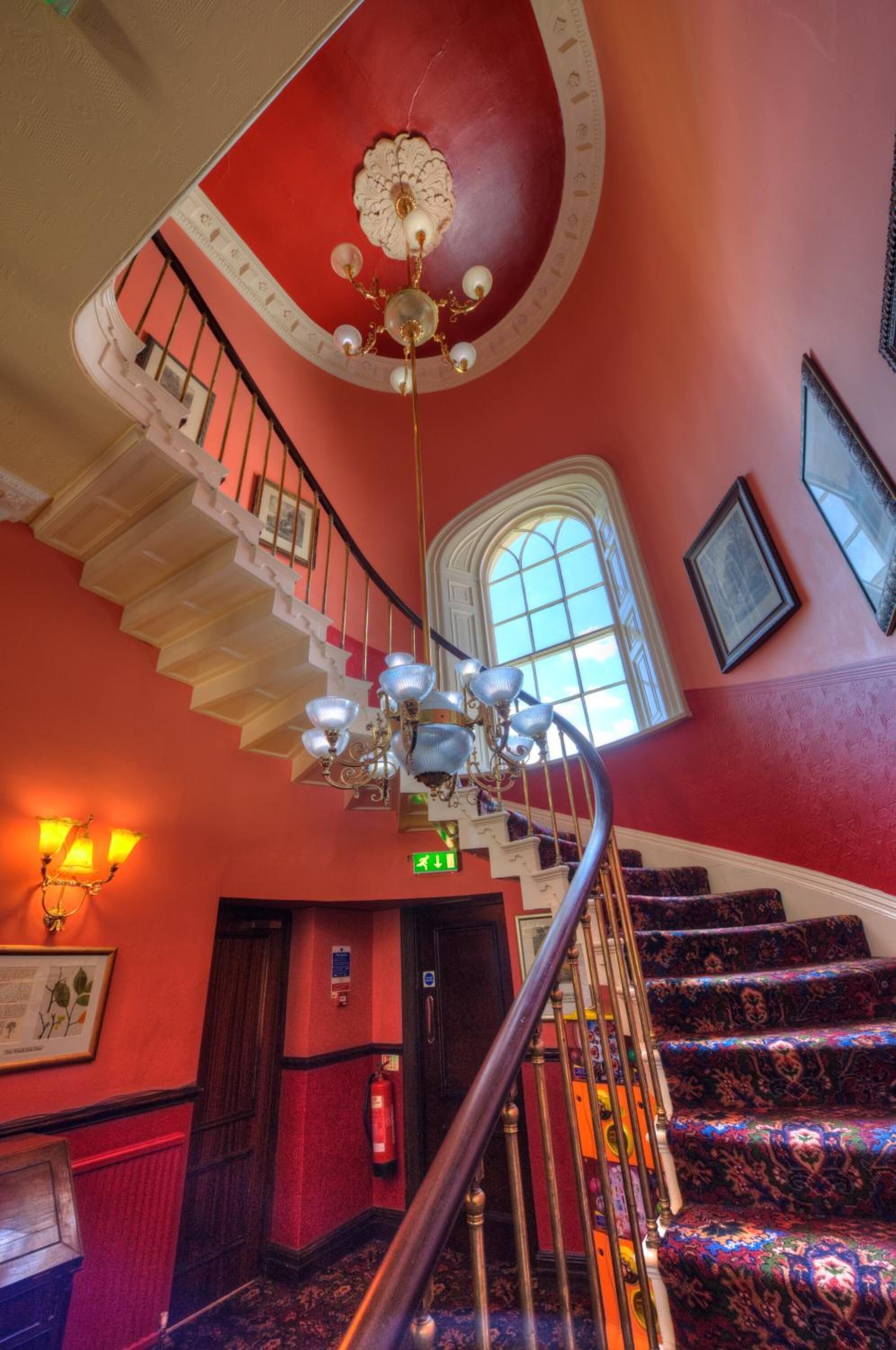 A grand staircase with ornate railings and a chandelier, leading to a landing with a red wall and a large window.