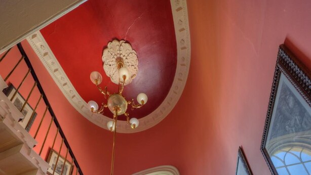 Een statige trap met sierlijke hekwerken en een gedessineerd tapijt, leidend naar een hoge plafond met een decoratieve kroonluchter.