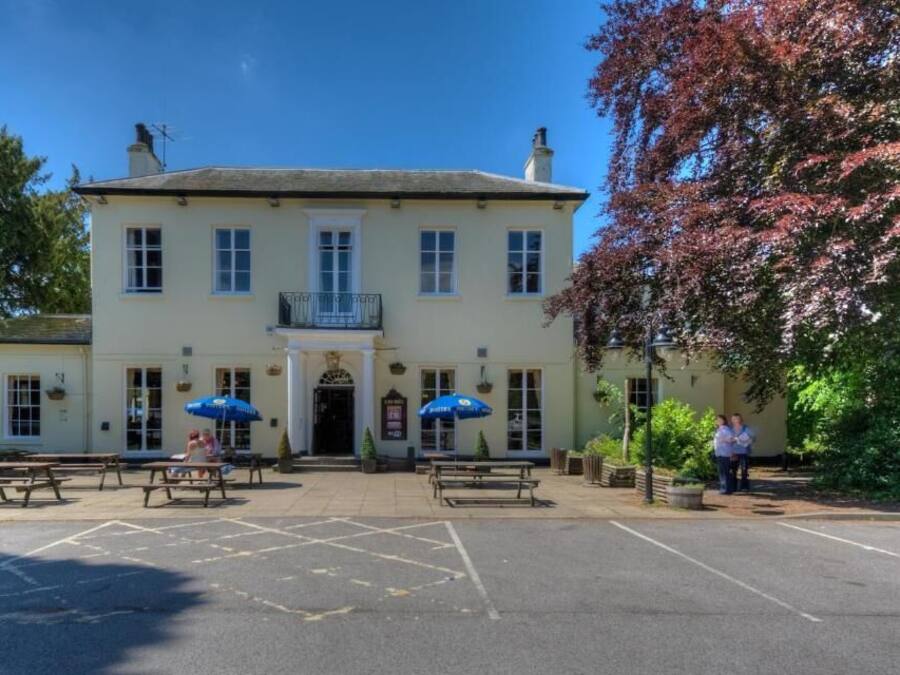 Un edificio de dos plantas con acceso central, zona de estar al aire libre y aparcamiento.