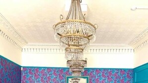 Een eetgedeelte met een grote kroonluchter, blauw bloemenbehang en gele stoelen.