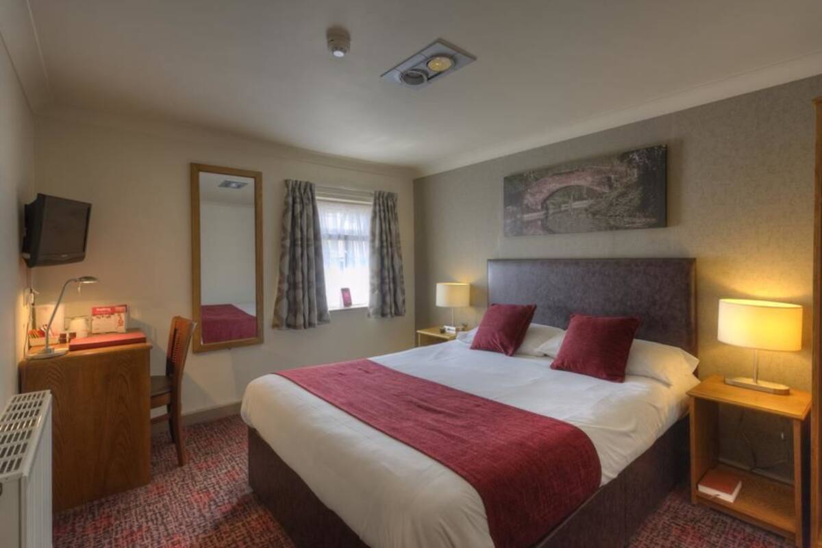 A hotel room with a large bed, two bedside tables with lamps, a desk with a chair, a television on the wall, and a window with curtains.