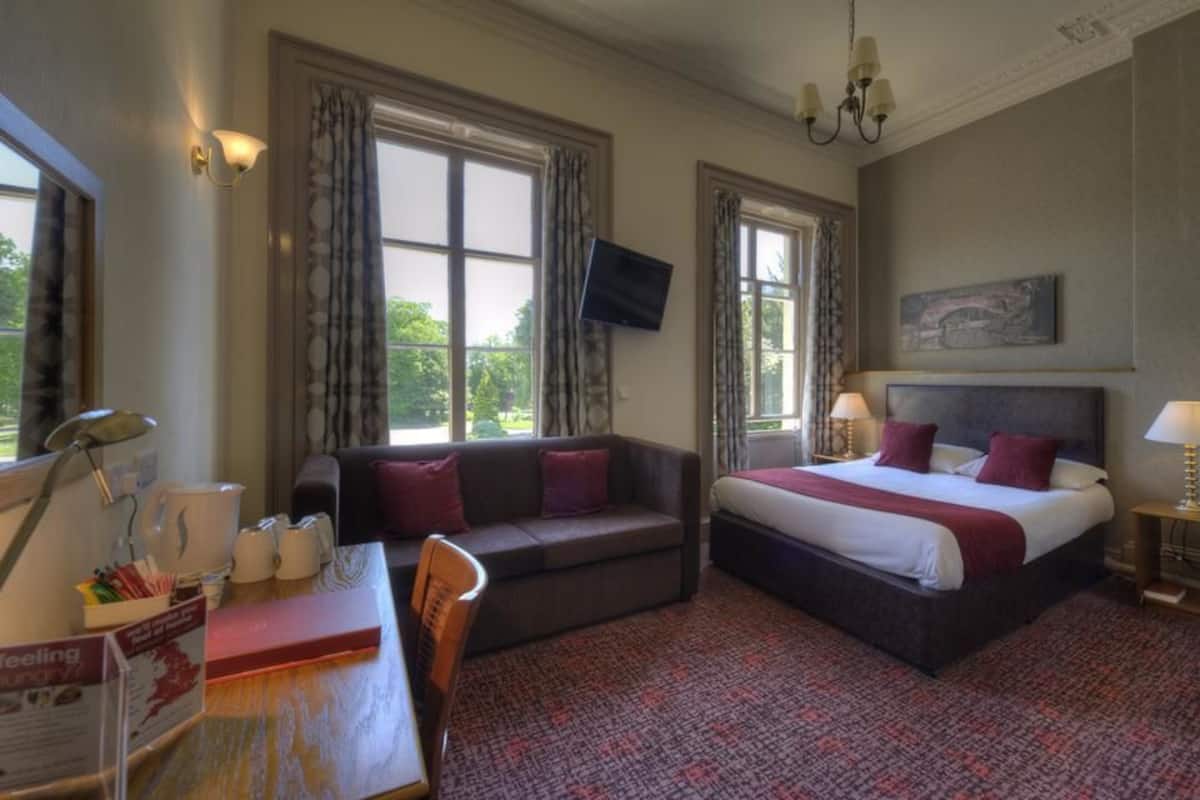 A hotel room with a bed, a sofa, a desk with a lamp, a television, and large windows with curtains.