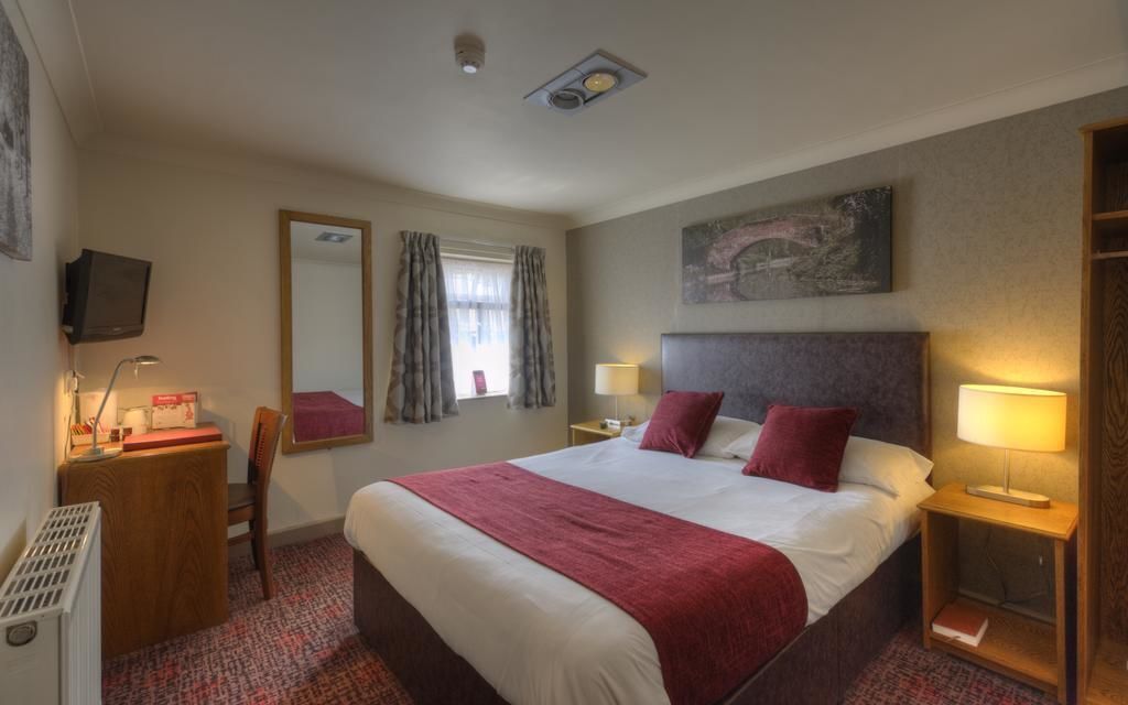 A hotel room with a large bed, bedside tables with lamps, a desk with a chair, a television, and a window with curtains.