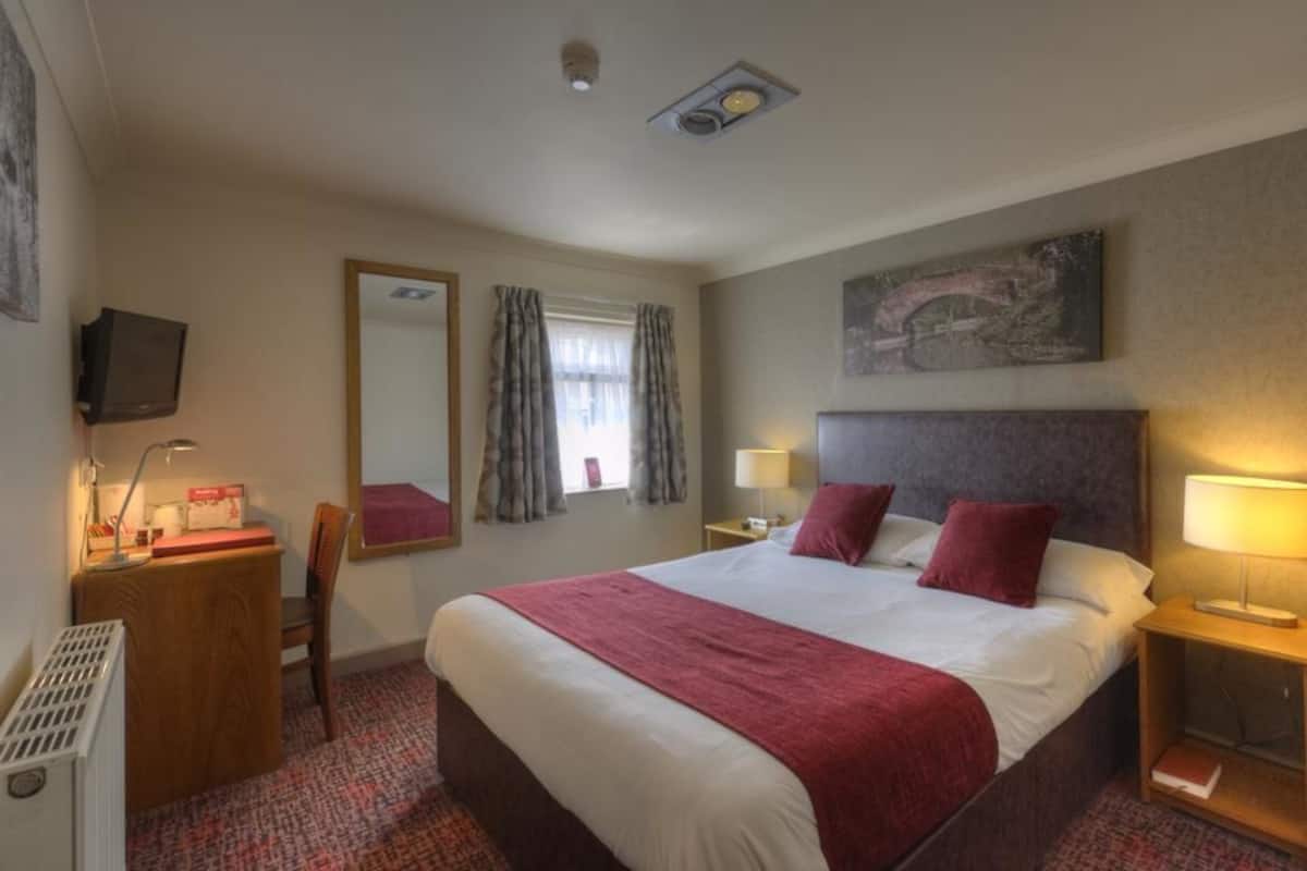 A hotel room with a large bed, bedside tables with lamps, a desk with a chair, a television, and a window with curtains.