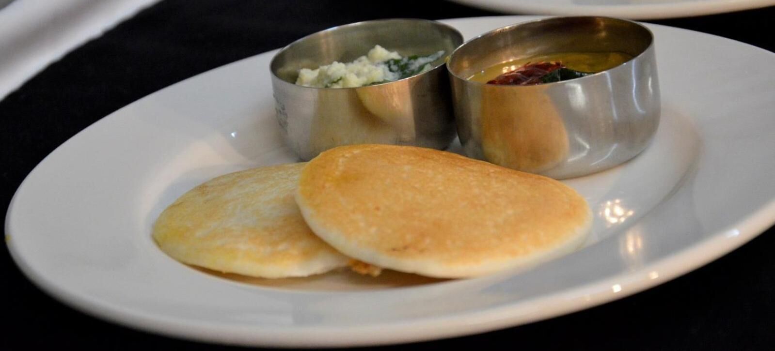 A plate with two pancakes and two small metal cups containing condiments.
