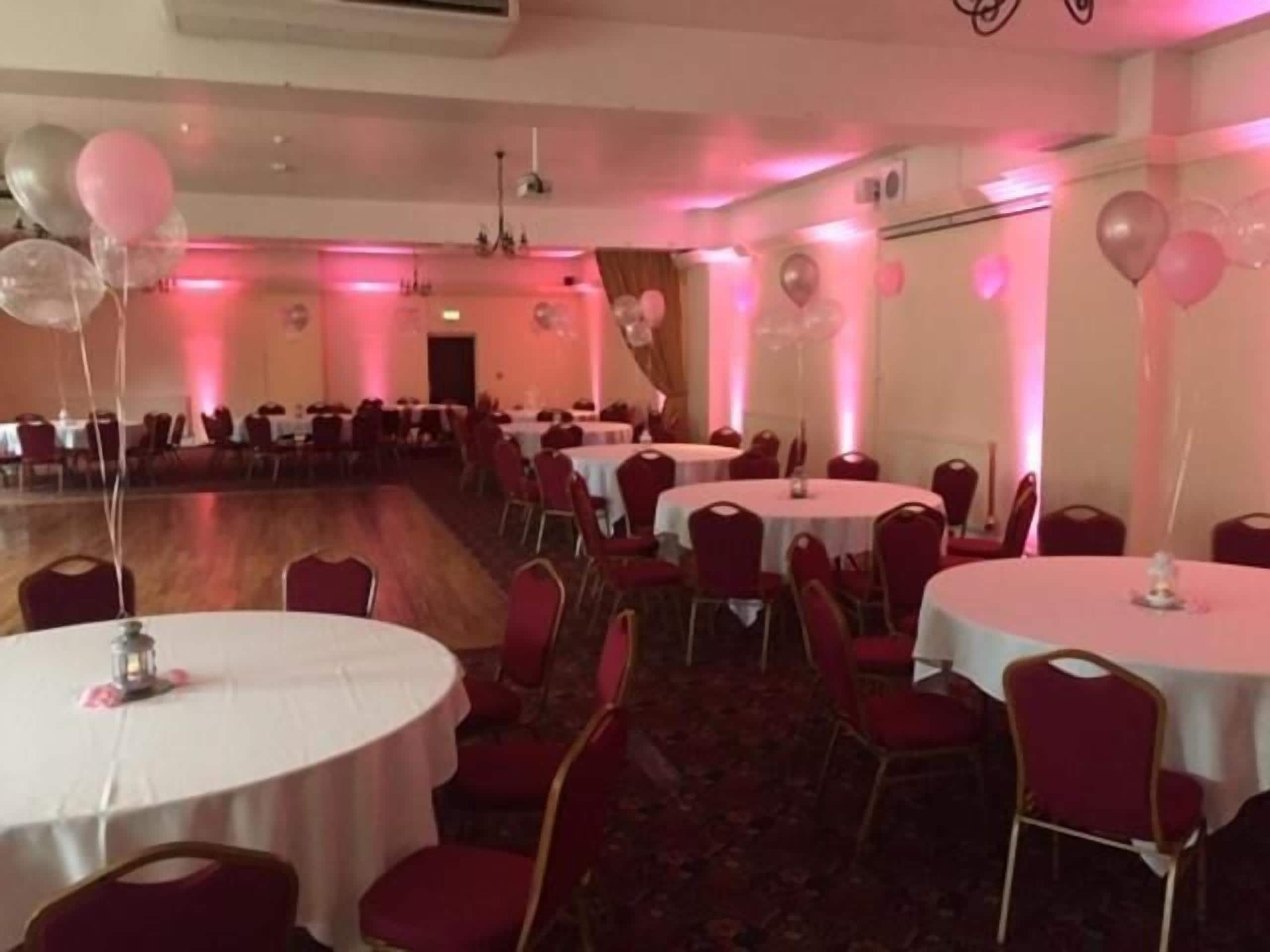 A banquet hall with round tables and red chairs, pink lighting, and balloons.