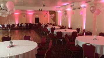 A banquet hall with round tables and red chairs, pink lighting, and balloons.