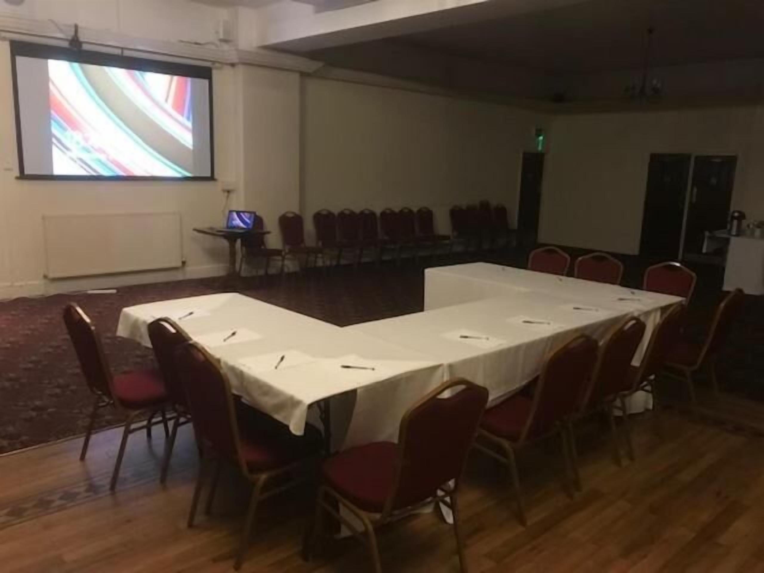A conference room with a long table, red chairs, a projector screen, and a laptop on a small table.