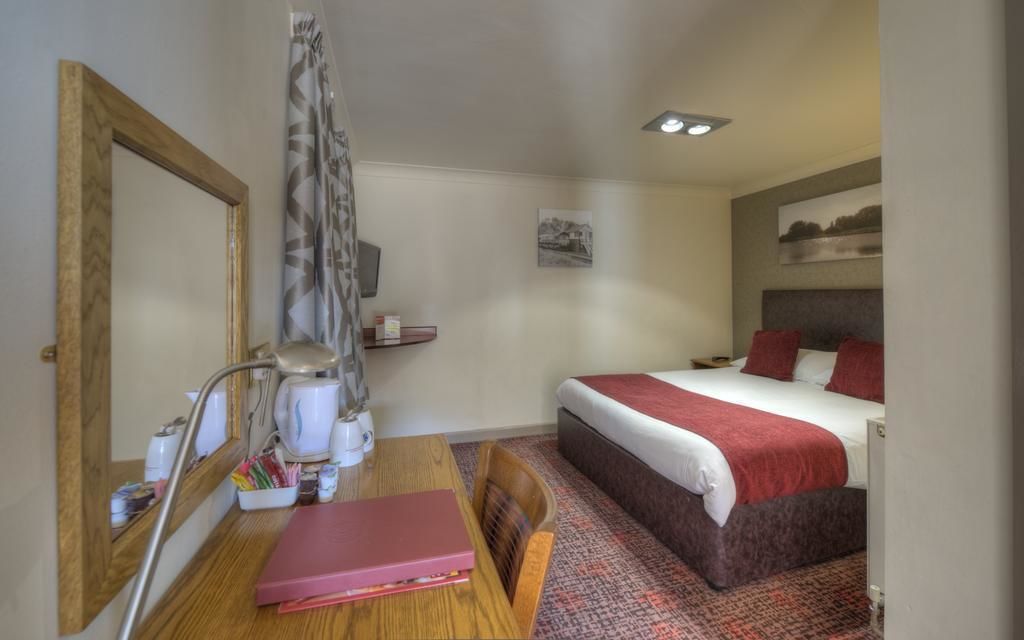 A compact hotel room with a single bed, a wooden desk with a laptop, a chair, a small table with a coffee maker, and a mirror.