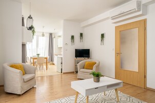 A modern living room with a sofa, a coffee table, and a TV. There is a wooden door and a white wall with hanging plants.