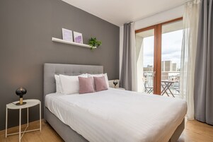 A modern bedroom with a large bed, a bedside table, a small shelf with pictures and a plant, and a view of the city through a large window.