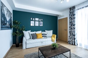 A modern living room with a teal accent wall, a white sofa with patterned cushions, a wooden coffee table, and a large window with geometric curtains.
