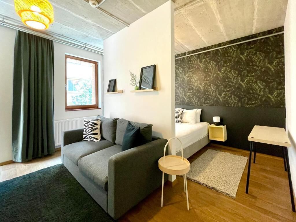 A modern living room with a sofa, a small table, a chair, a bed, and a window with curtains.