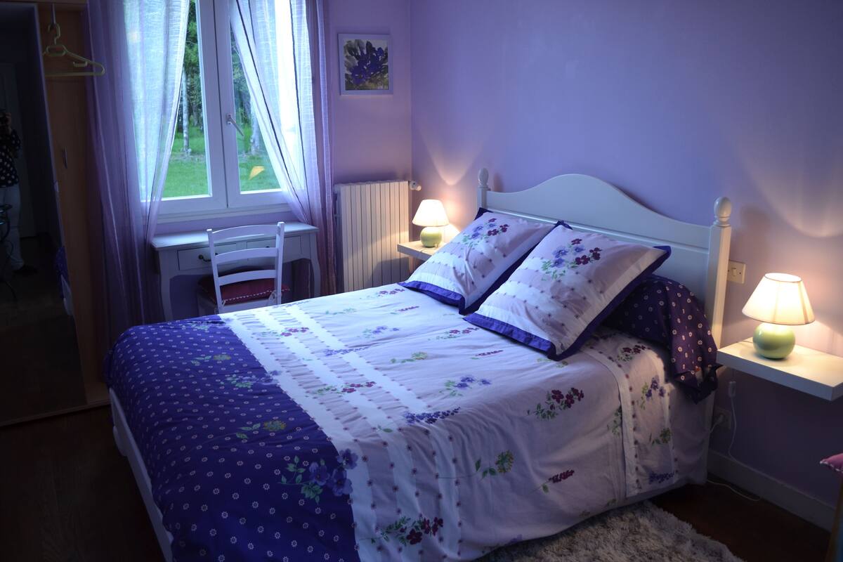 A bedroom with a bed, two bedside lamps, a window with curtains, a radiator, and a desk with a chair.