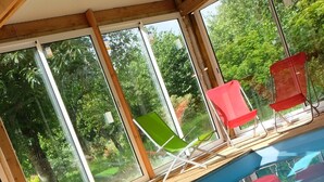An indoor swimming pool with clear glass walls, wooden deck, and red and green lounge chairs. - Chambres d'Hôtes La Semondière (Saint-Thomas-de-Courceriers)