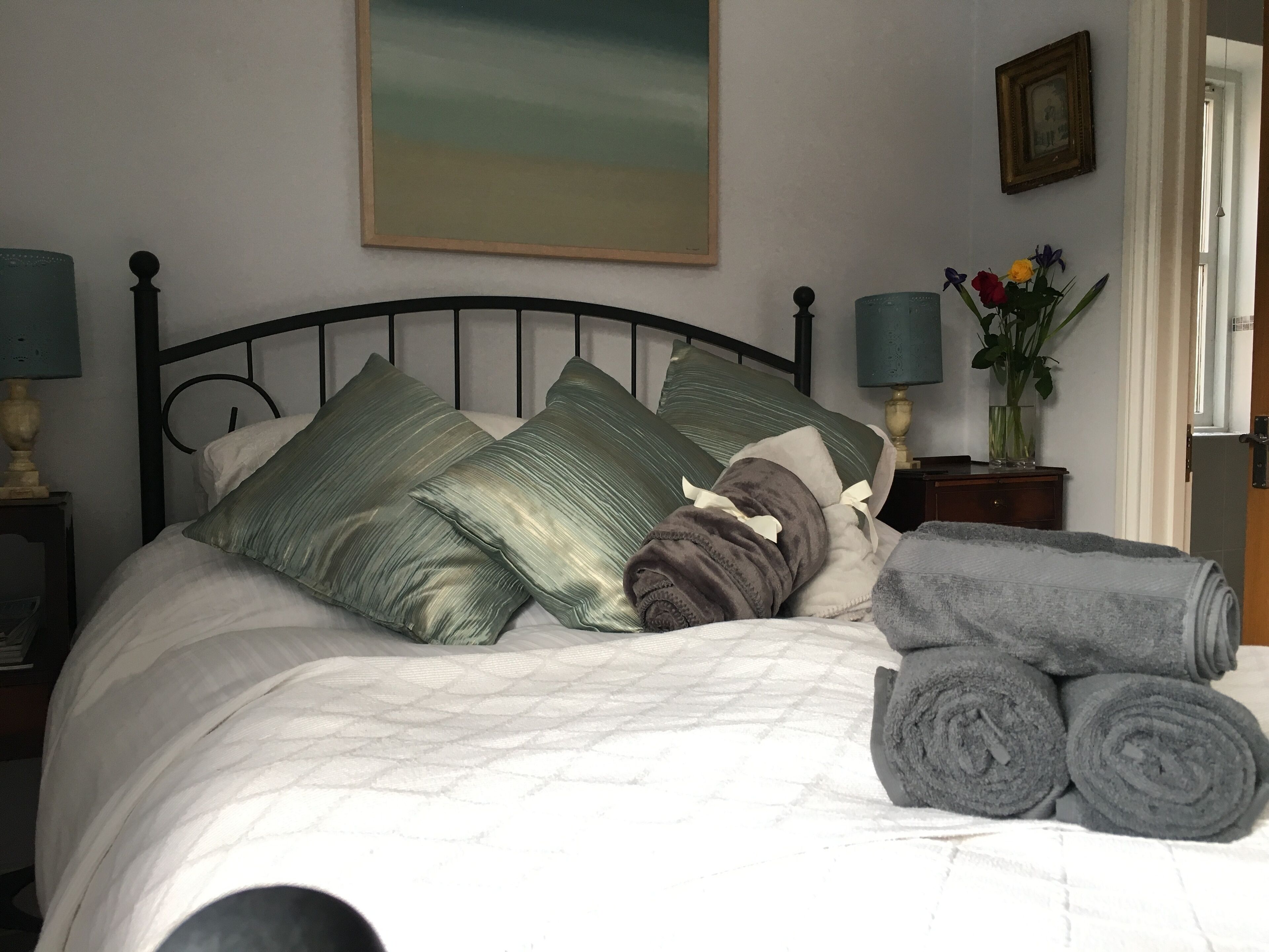 Una cama bien hecha con almohadas y toallas enrolladas, un cuadro en la pared y un florero con flores sobre una mesita de noche.