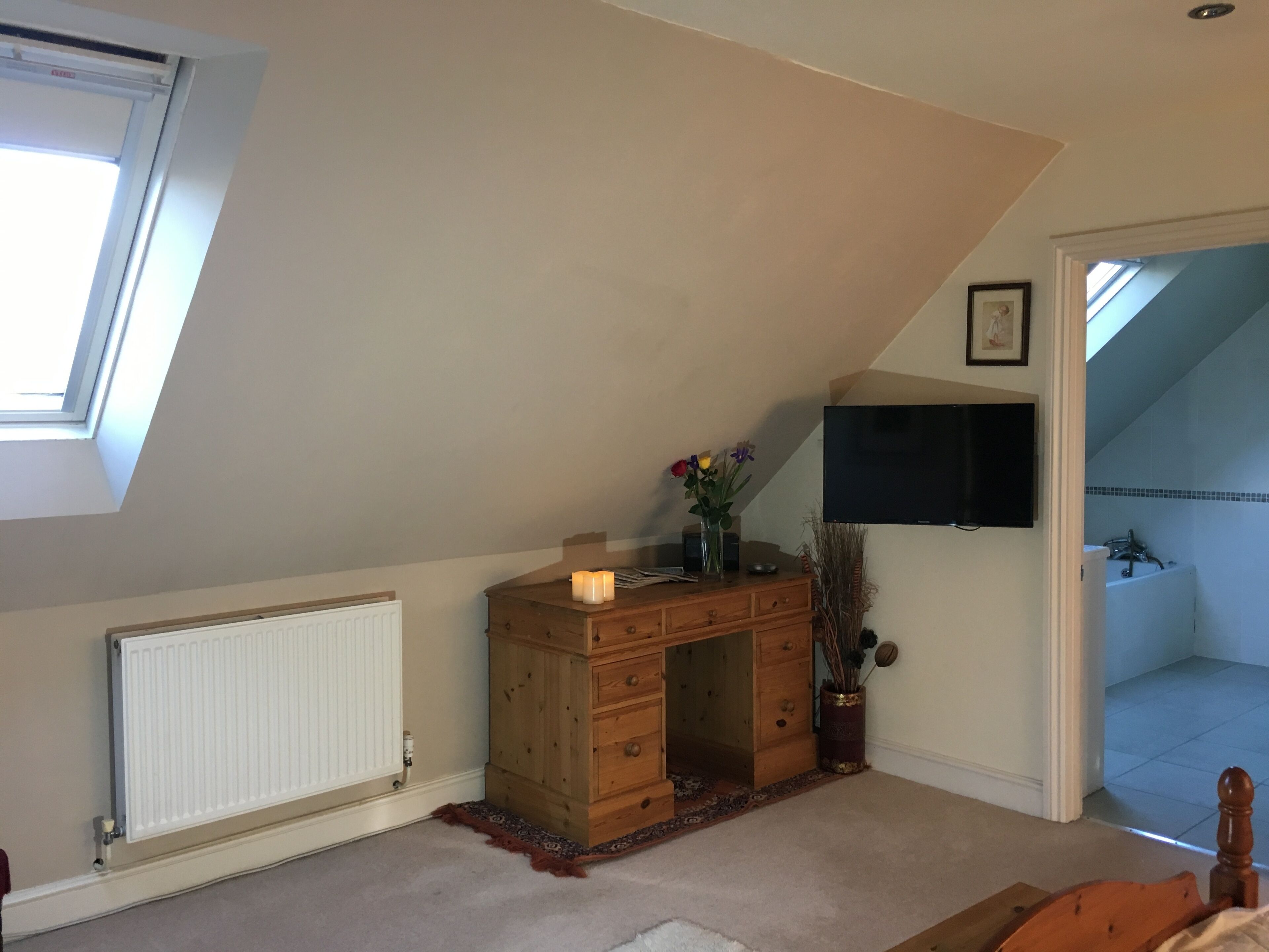 Una habitación con techo inclinado, un aparador de madera, un radiador, un televisor montado en la pared y una puerta que da a otra habitación.
