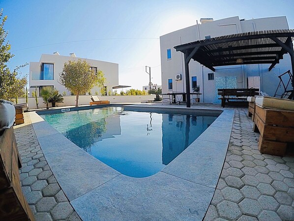 A swimming pool with a hexagonal tiled edge, surrounded by a patio area with hexagonal tiles. There are wooden benches and a pergola structure. - Villa Amor (Rhodes)
