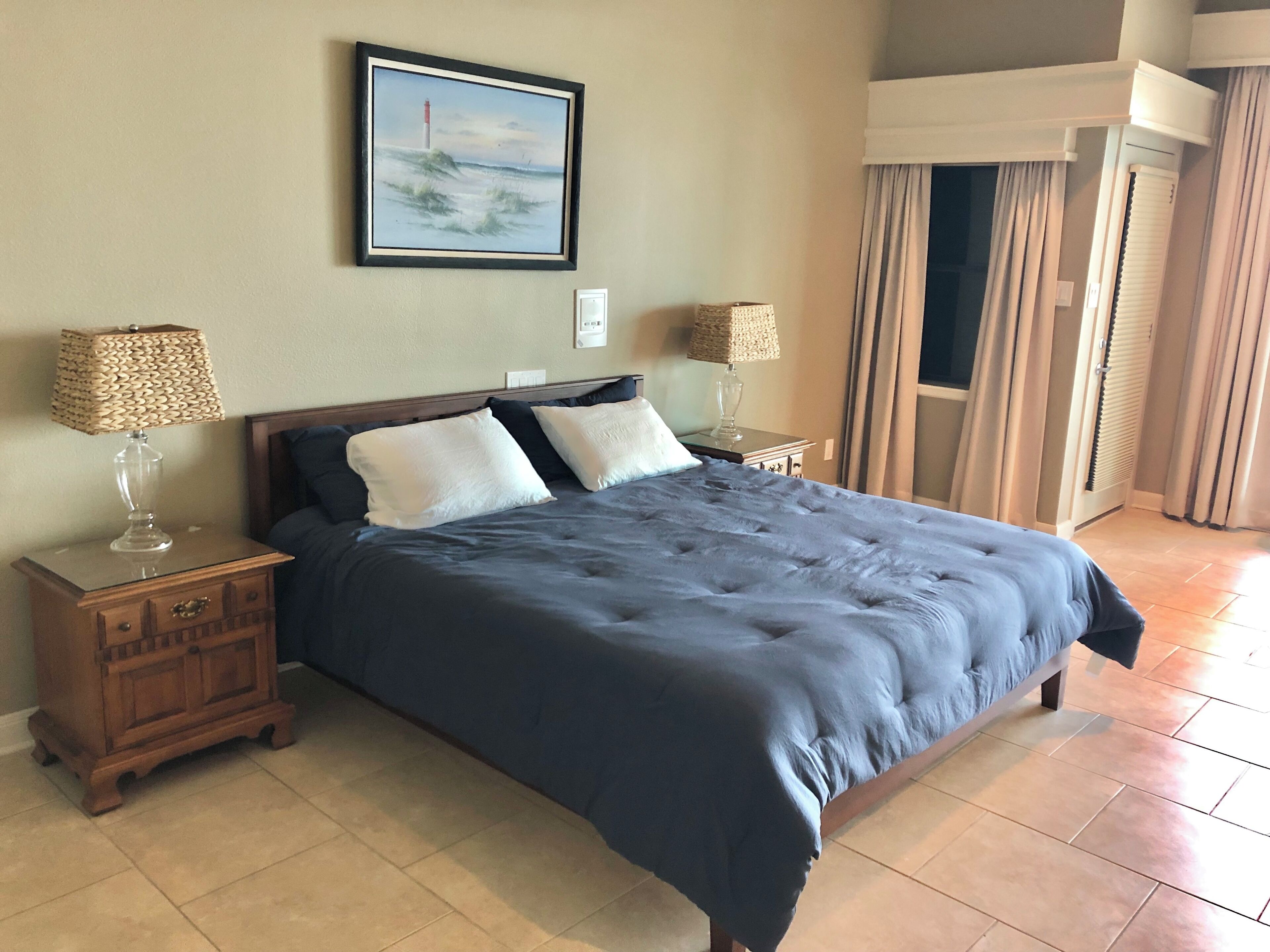 A neatly made bed with a dark blue duvet, two bedside tables with lamps, a framed painting of a lighthouse, and a view of a balcony with curtains.