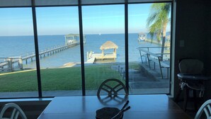 A view of the ocean through large windows, a wooden table with a bowl and spoon, and a chair. - The Bay House Kemah - Houston area Bayfront Home w/ Elevator & Bay Views (Kemah)