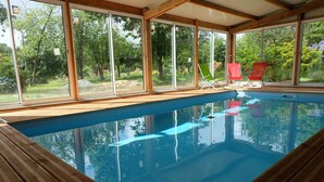 Una piscina interior con paredes de vidrio transparente, terraza de madera y sillones reclinables rojos.