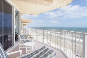 Un balcón con vistas a la playa, muebles de exterior blancos y una puerta de vidrio.