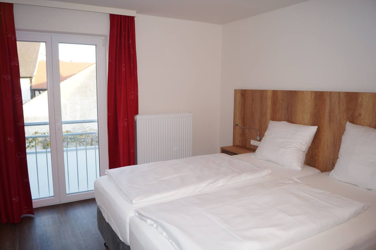 A hotel room with a double bed, a wooden headboard, a window with red curtains, and a view of a building outside.