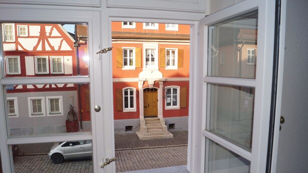 Vista a través de una puerta de un edificio con fachada roja, una escalera de madera que conduce a una puerta y un coche estacionado al frente.