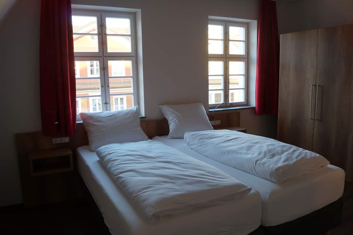 A double bed with white linens in a room with two windows and a wooden wardrobe.