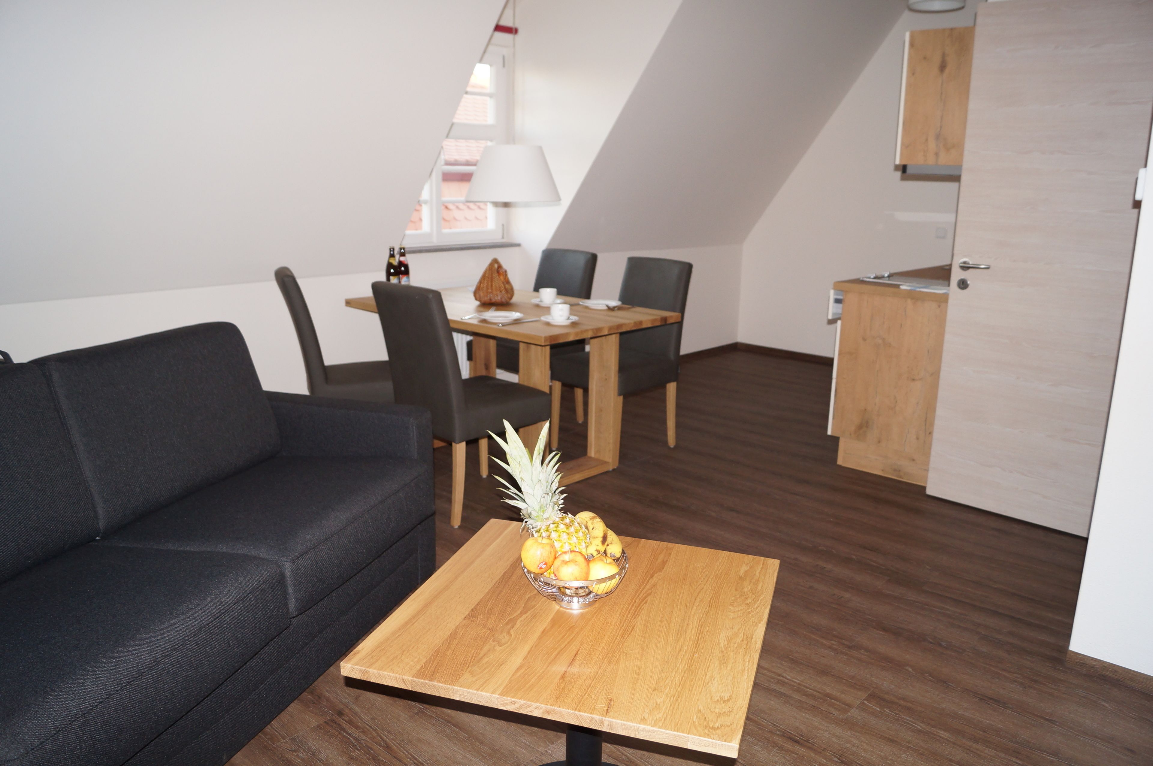 A modern living room with a dark grey sofa, a wooden dining table with chairs, and a fruit bowl on a small table.