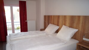 Una cama doble con sábanas blancas en una habitación con cabecera de madera, una mesita de noche y vistas a un balcón con cortinas rojas.