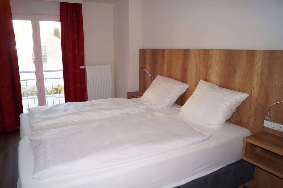 A double bed with white linens in a room with a wooden headboard, a bedside table, and a view of a balcony with red curtains.